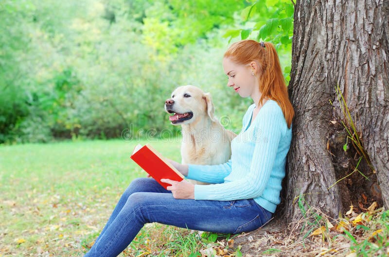 Happy owner woman reading book with Golden Retriever dog sitting near tree in park