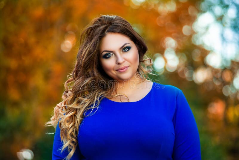 Happy plus size model in blue dress outdoors, fat woman in autumn park among yellow leaves