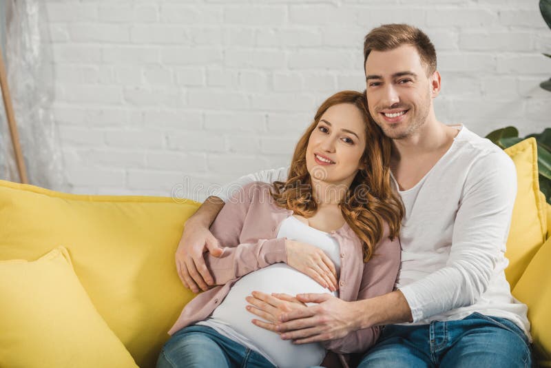 happy pregnant couple sitting on couch and smiling stock images