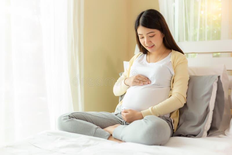 Happy pregnant young asian woman touching belly and admiring her baby or fetus on bed with happiness smile face. Young Asia mother