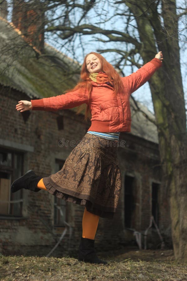 A happy red-haired young girl in a bright red jacket and pantyhose, in a skirt with floral patterns, poses against the backdrop of nature and the old village house