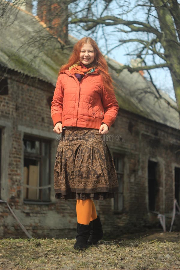 A happy red-haired young girl in a bright red jacket and pantyhose, in a skirt with floral patterns, poses against the backdrop of nature and the old village house