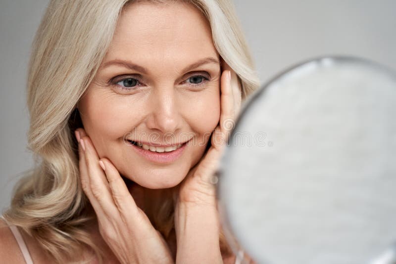 Happy 50s middle aged woman touching face skin looking in mirror.