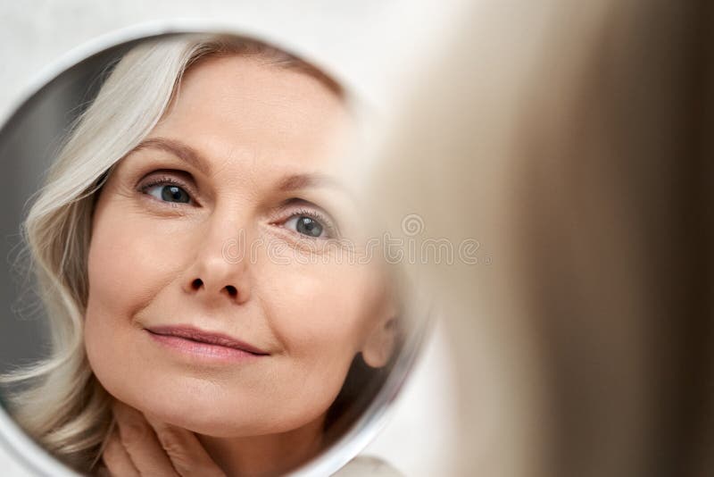 Happy 50s middle aged woman touching face skin looking in mirror reflection.