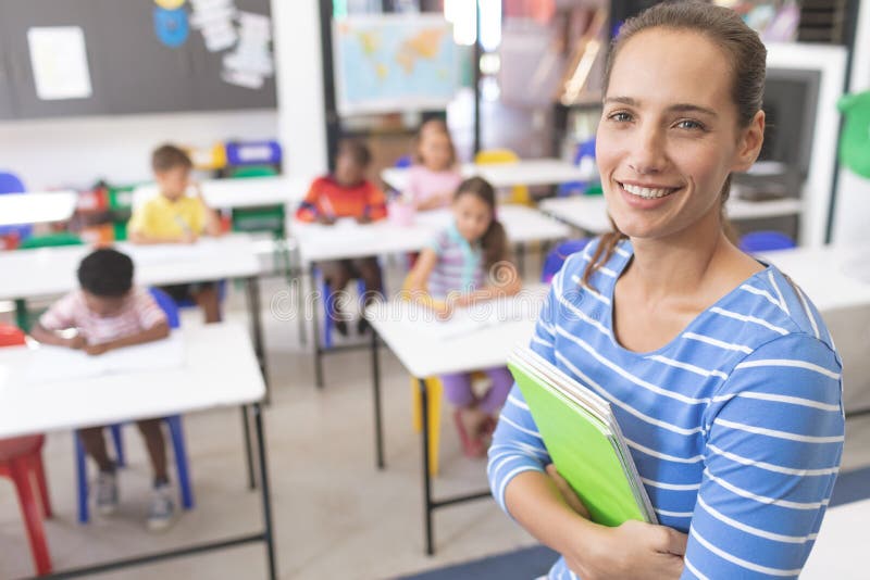 Side view of happy schoolteacher standing in classroom and holding a book against pupils sitting on their chair in background. Side view of happy schoolteacher standing in classroom and holding a book against pupils sitting on their chair in background