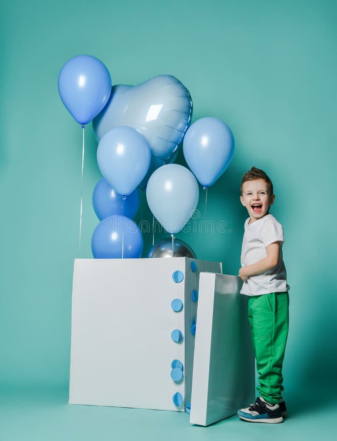 Happy screaming kid boy stands at white box he has just opened with blue balloons for birthday party flying out of it