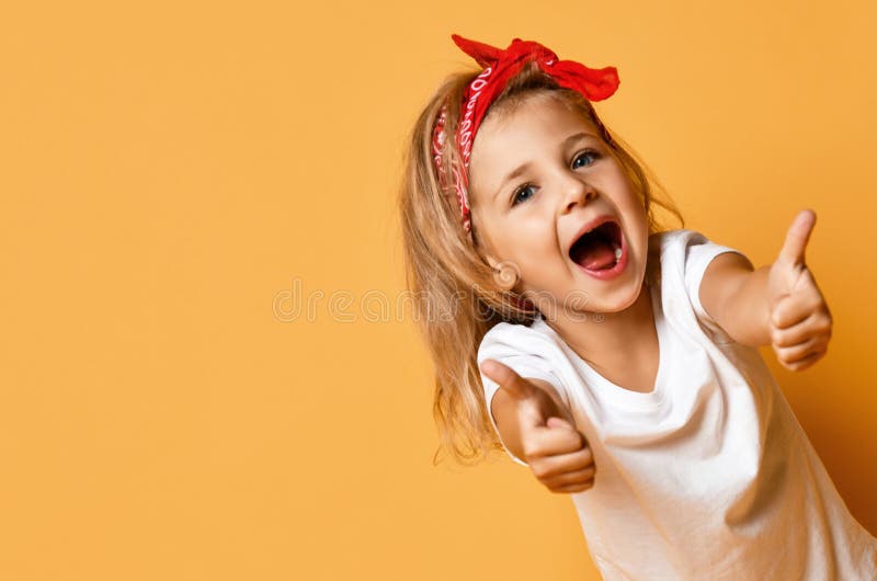 Happy screaming kid girl in white t-shirt and red headband is showing thumbs up gesture at free copy space on yellow