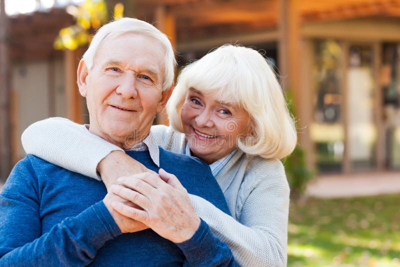 Happy senior couple bonding to each other and smiling while standing outdoors and in front of their house. Happy senior couple bonding to each other and smiling while standing outdoors and in front of their house