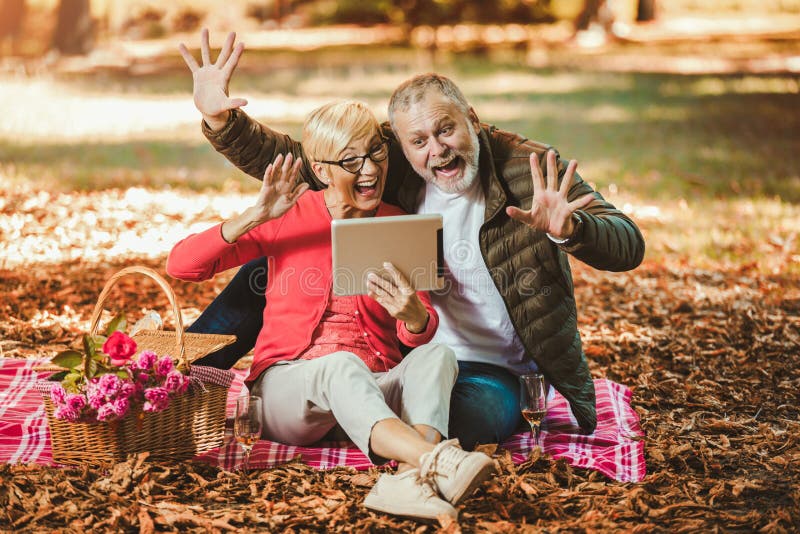 Senior couple having a picnic in autumn park make video call using digital tablet