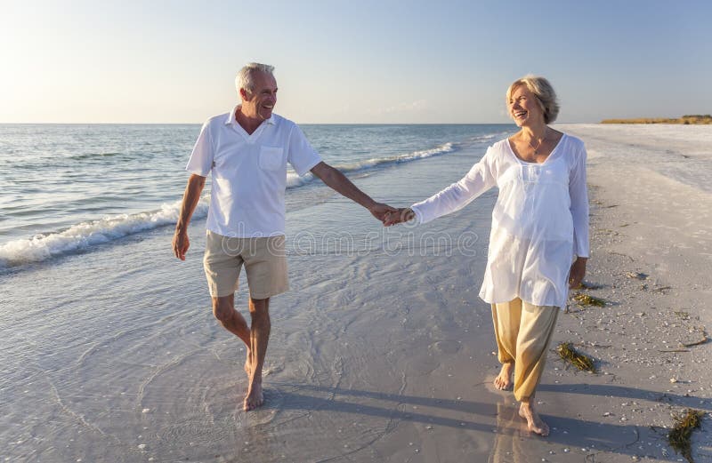 Felice senior, uomini e donne, coppia a piedi e tenendo le mani su una deserta spiaggia tropicale con un luminoso cielo blu chiaro.