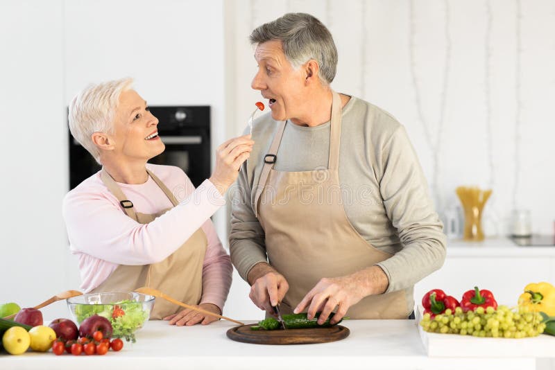 Happy Senior Wife Feeding Husband Cooking Together Standing In Kitchen