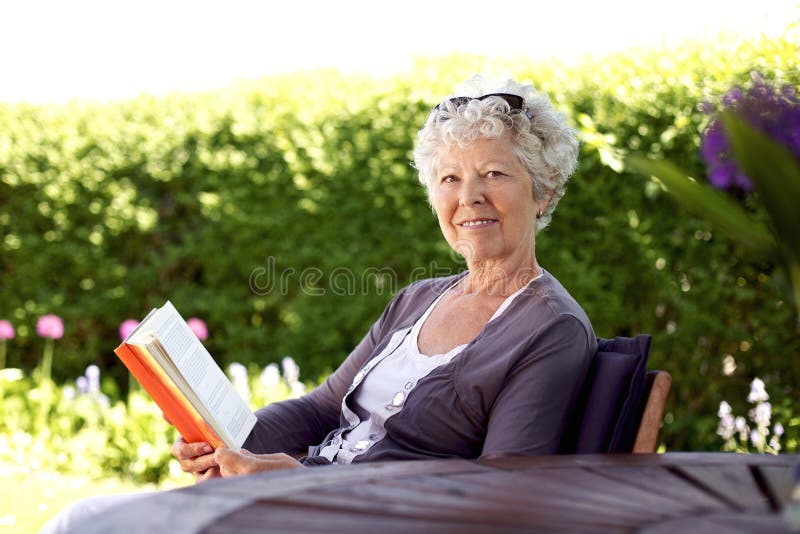 Happy senior woman with book in hand sitting in her backyard looking at camera smiling - Elder woman reading novel in garden. Happy senior woman with book in hand sitting in her backyard looking at camera smiling - Elder woman reading novel in garden