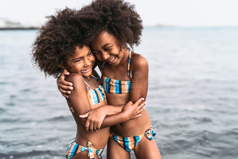 Happy sisters embracing inside sea water during summer time - Afro kids having fun playing on the beach