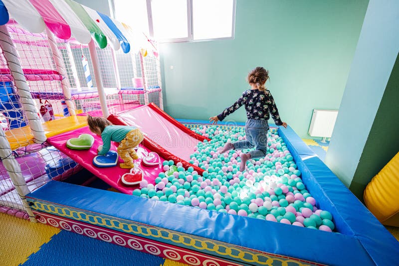 Happy sisters playing at indoor play center playground. Girl jump at color balls in ball pool