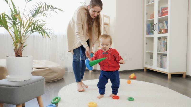 Happy smiling baby boy holding toys and learning walking on carpet in living room. Baby development, family playing games, making