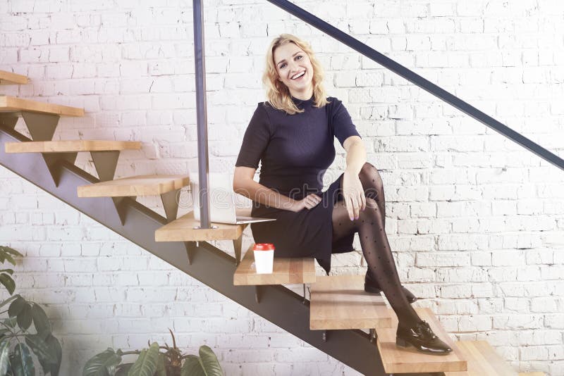 Happy smiling businesswoman sitting on stairs in modern office, working at laptop and having coffee, sunshine daylight.