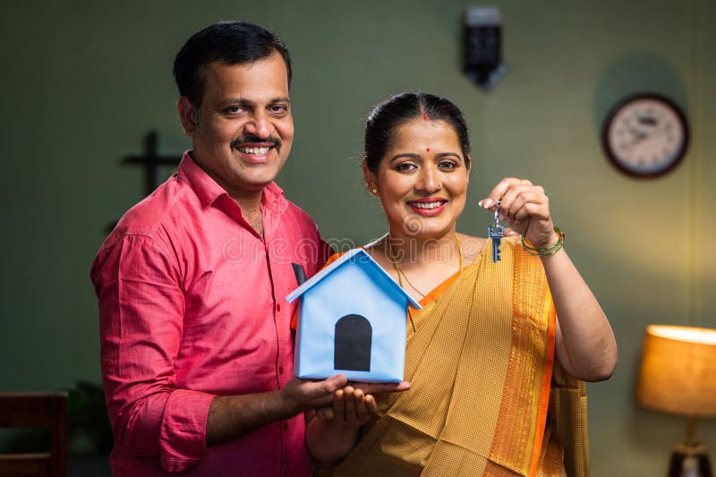 Happy smiling couple showing new house keys by toy home while looking camera - concept of new home purchasing, home loan