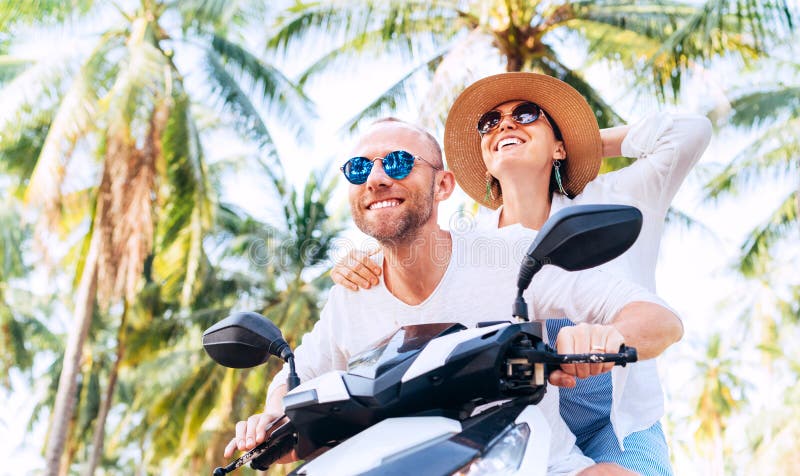 Happy smiling couple travelers riding motorbike during their tropical vacation under palm trees