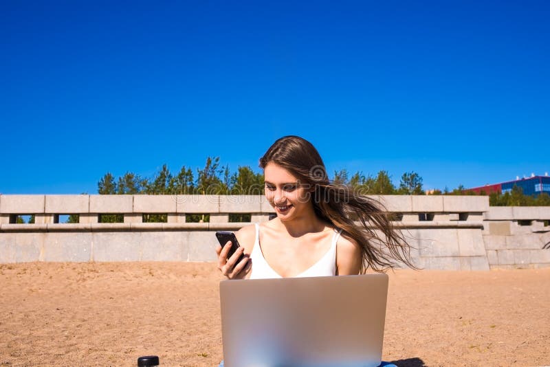 Happy smiling female copywriter watching funny video on mobile phone during distance work on laptop computer, sitting on the beach stock photography Happy smiling female copywriter watching funny video on mobile phone during distance work on laptop computer, sitting on the beach stock photography