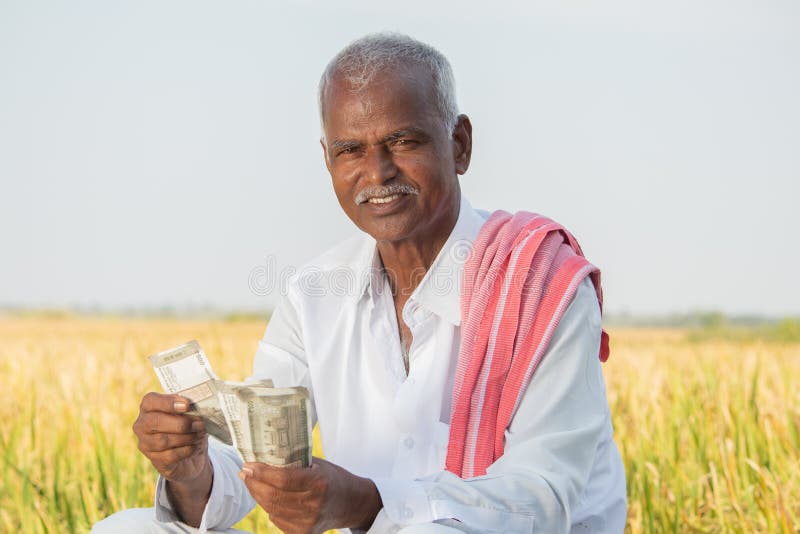 Happy smiling Indian farmer counting money on agriculture field while looking camera - concept of good or bumper crop harvest