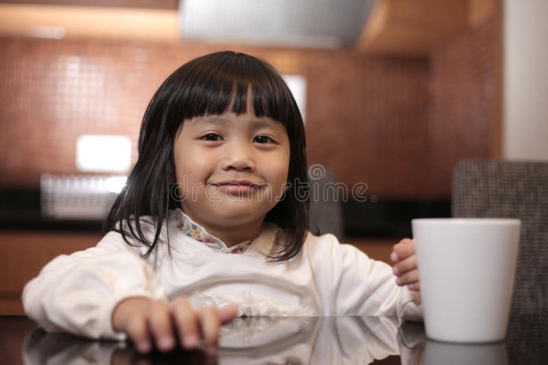 Happy smiling little Asian girl drinking a glass of milk or tea in white cup stock photography