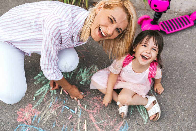 Happy smiling little girl and mother laughing and drawing with chalks on the sidewalk. Caucasian female play together with her cute kid preschooler with backpack outdoor. Mom and child activity