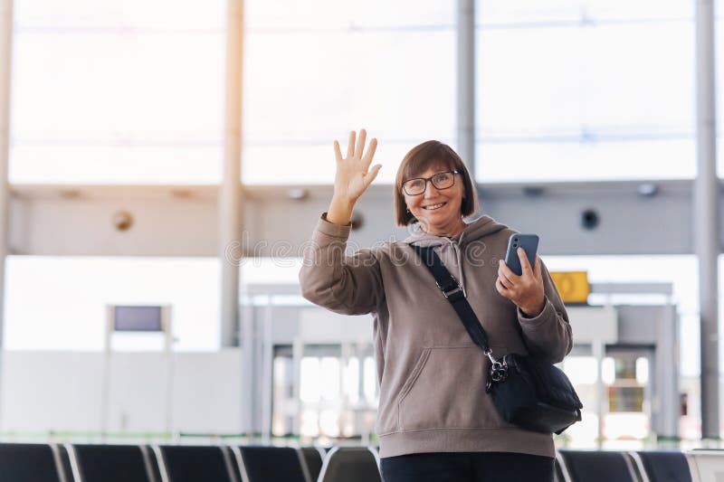 Happy smiling mature traveler woman hold smartphone and wave hand in greeting someone who meeting her at airport