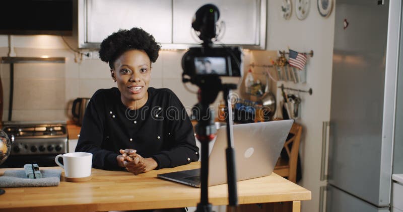 Happy smiling young beautiful black vlogger woman filming new video using professional camera at home table slow motion. royalty free stock photo