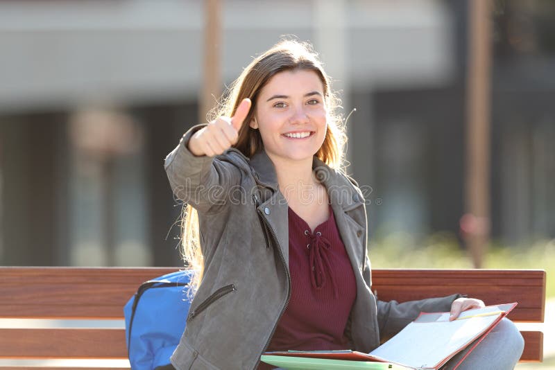 Happy student gesturing thumb up looking at camera sitting on a bench in a park. Happy student gesturing thumb up looking at camera sitting on a bench in a park