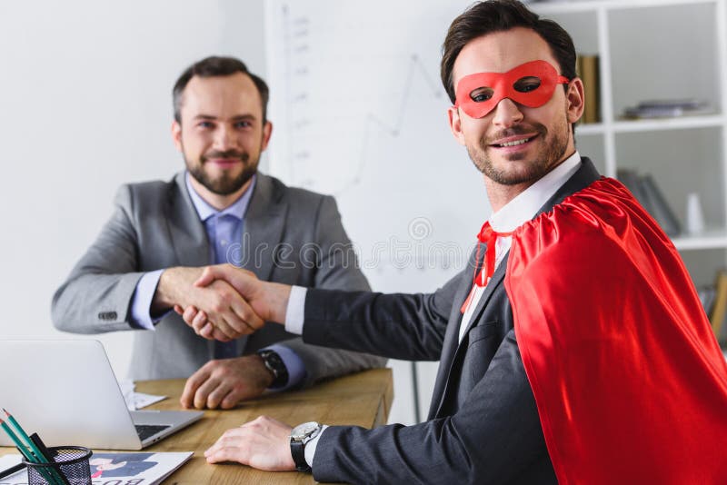 happy super businessman in mask and cape shaking hands with businessman stock photo happy super businessman in mask and cape shaking hands with businessman stock photo