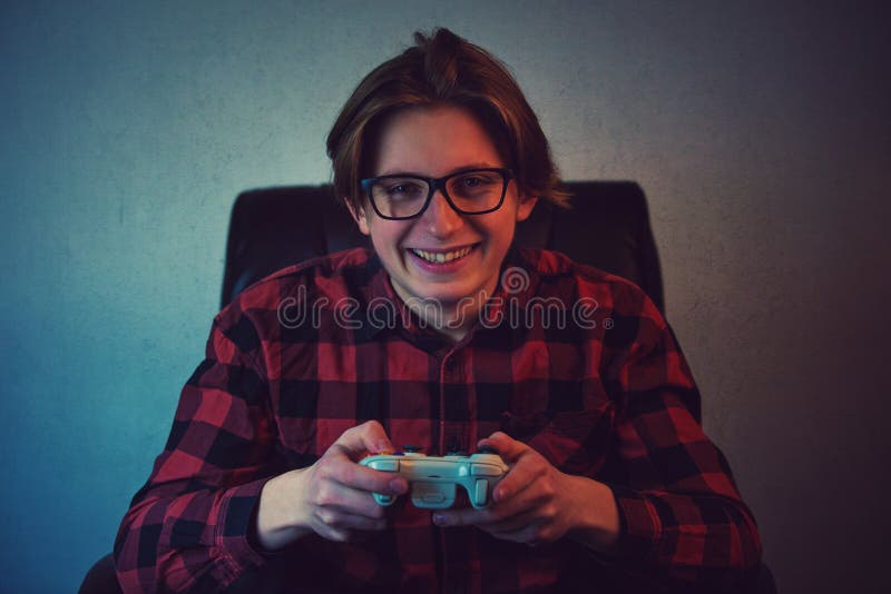 Happy teenage boy playing video games seated in his armchair. Excited adolescent wearing eyeglasses, holding joystick console royalty free stock photography Happy teenage boy playing video games seated in his armchair. Excited adolescent wearing eyeglasses, holding joystick console royalty free stock photography