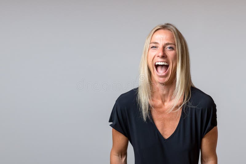 Happy temperamental mature blond woman yelling and glaring at the camera with her mouth wide open isolated on grey with copy space. Happy temperamental mature blond woman yelling and glaring at the camera with her mouth wide open isolated on grey with copy space