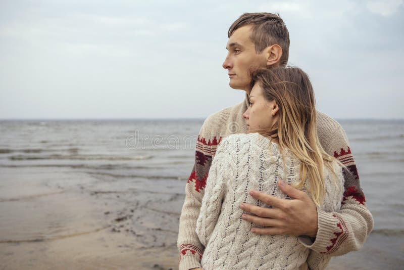 Happy thoughtful couple standing on a rock beach near sea hugging each other in cold foggy cloudy autumn weather. Copy space