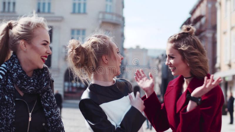 Happy three friends wandering in the old city center, laughing, and joking. The woman in a red coat hugs her friend