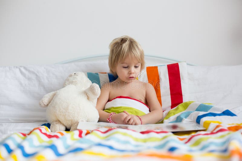 Happy toddler child, blond boy, sitting in bed with stuffed toy after bath, reading a book