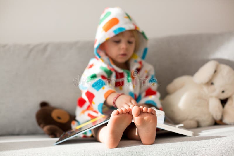 Happy toddler child, blond boy, sitting on the couch with stuffed toy after bath, reading a book