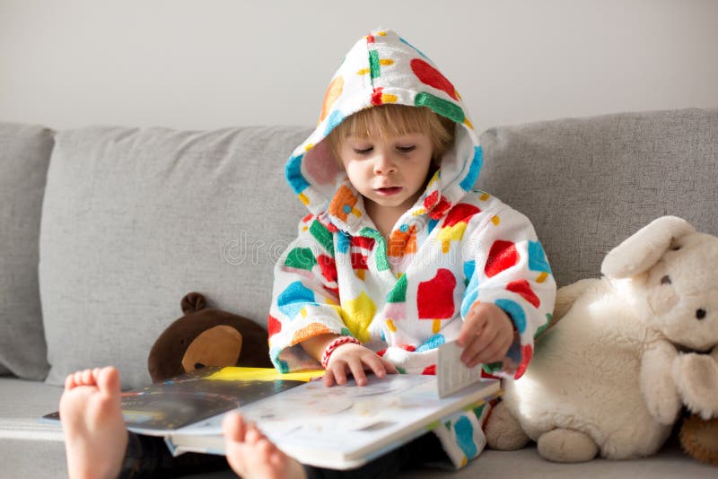 Happy toddler child, blond boy, sitting on the couch with stuffed toy after bath, reading a book