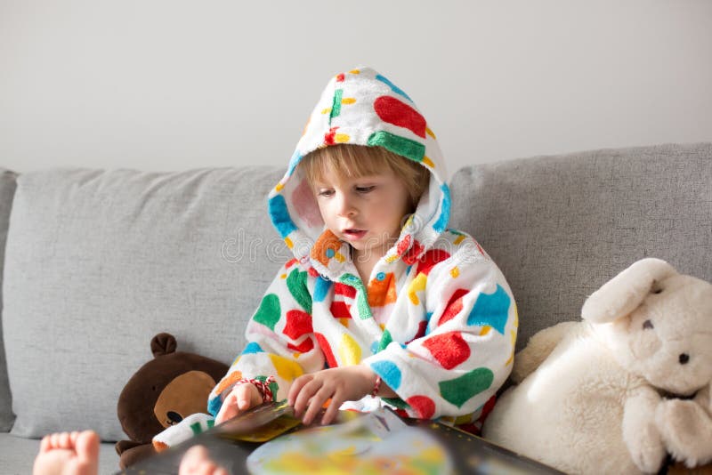 Happy toddler child, blond boy, sitting on the couch with stuffed toy after bath, reading a book