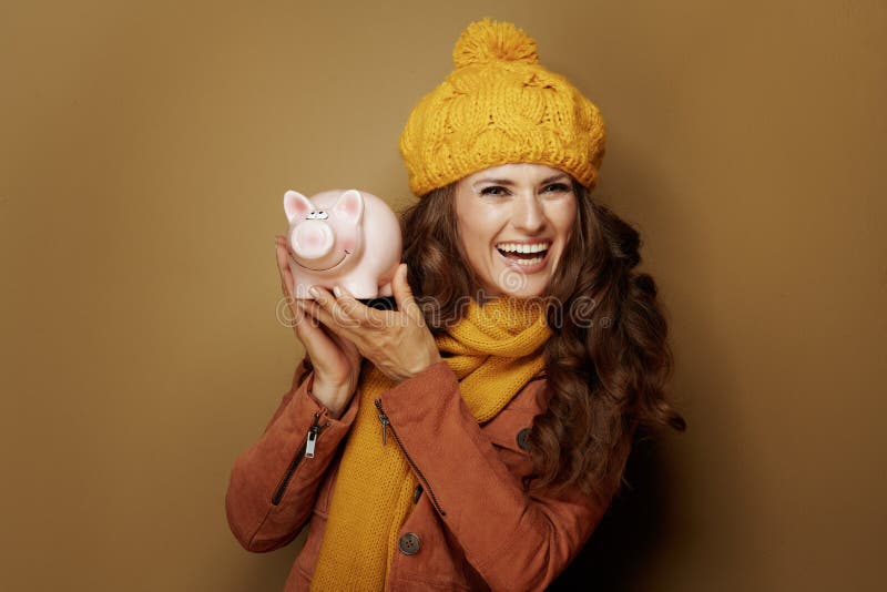 Happy trendy woman in yellow beret and scarf showing piggy bank on bronze background. Happy trendy woman in yellow beret and scarf showing piggy bank on bronze background