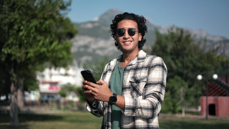 Happy Turkish man walking at summer sunny city park scenery chatting use smartphone relaxing outdoor stock footage