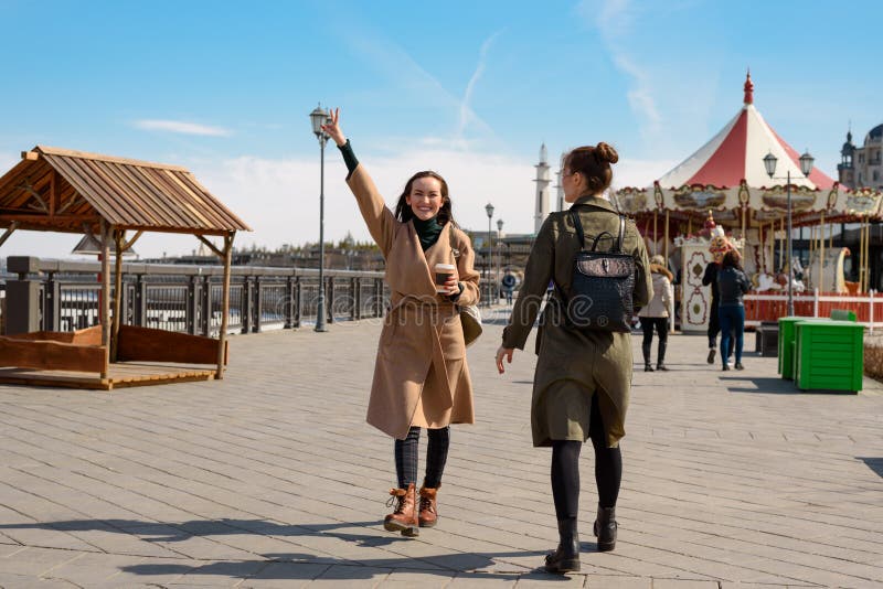 Happy two female students of a friend walk around the city in stylish coats and backpacks and drink coffee from disposable cups, a royalty free stock image Happy two female students of a friend walk around the city in stylish coats and backpacks and drink coffee from disposable cups, a royalty free stock image