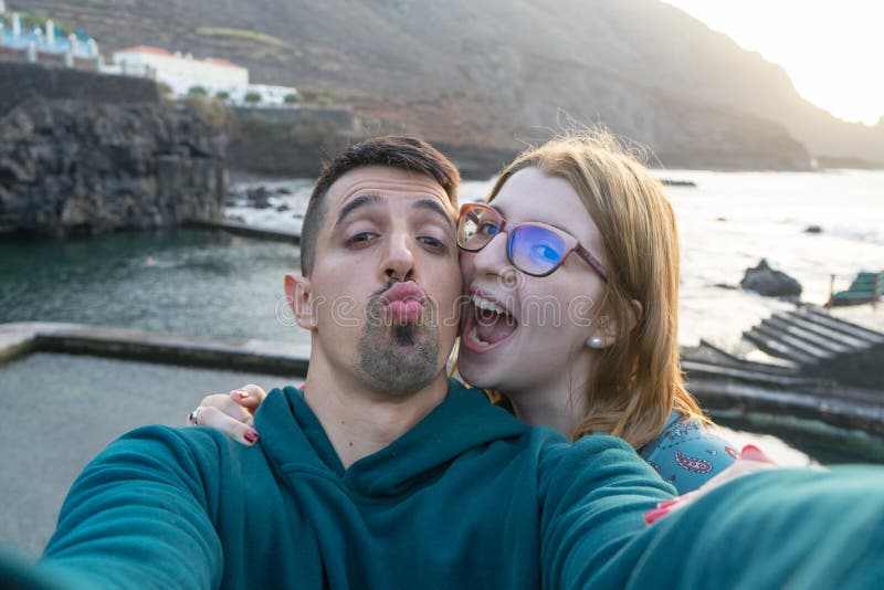 Happy wife with glasses laughing, giving hug with tenderness her latin husband, sending kiss to camera, flirting in coast. Friends