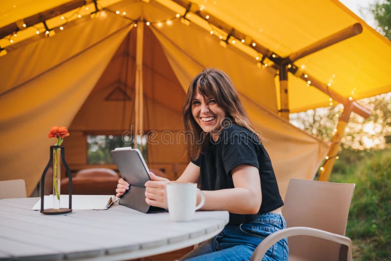 Happy Woman freelancer using a laptop on a cozy glamping tent in a sunny day. Luxury camping tent for outdoor summer holiday and