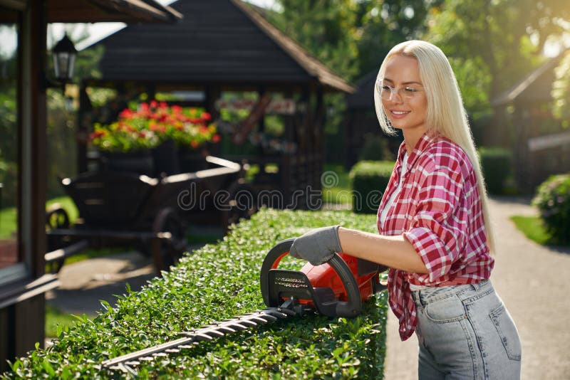 Happy caucasian woman in safety glasses and gloves shaping bushes with electric trimming machine. Concept of people, gardening and seasonal work. Happy caucasian woman in safety glasses and gloves shaping bushes with electric trimming machine. Concept of people, gardening and seasonal work.