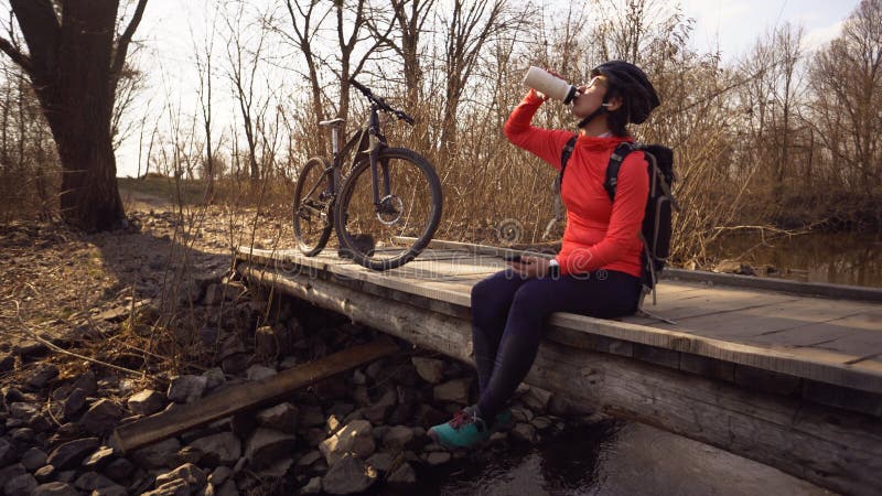 Happy woman sits wooden bridge over river, listening music in headphones with smartphone hands and drinks water white bottle. stock photos Happy woman sits wooden bridge over river, listening music in headphones with smartphone hands and drinks water white bottle. stock photos