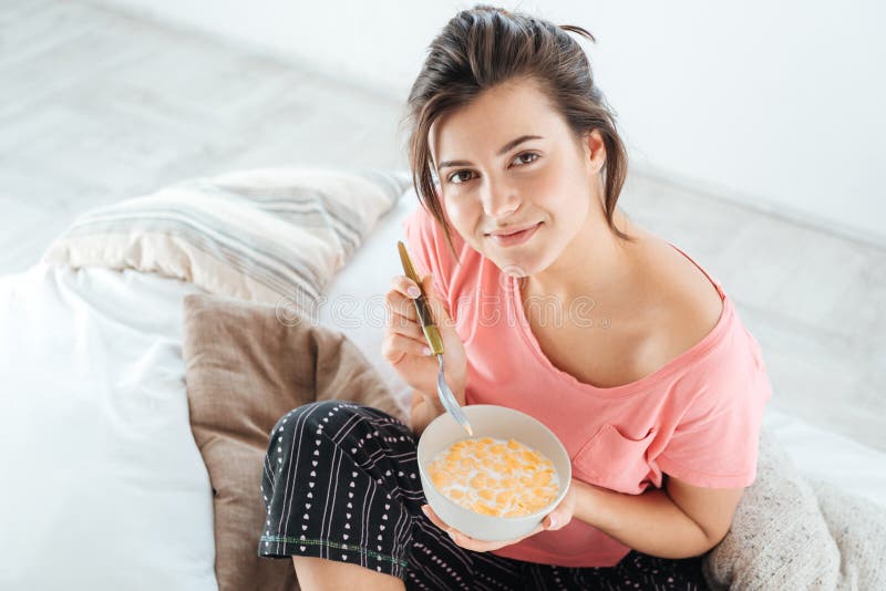 Top view of happy lovely young woman in pajamas sitting on bed and eating cereals with milk. Top view of happy lovely young woman in pajamas sitting on bed and eating cereals with milk