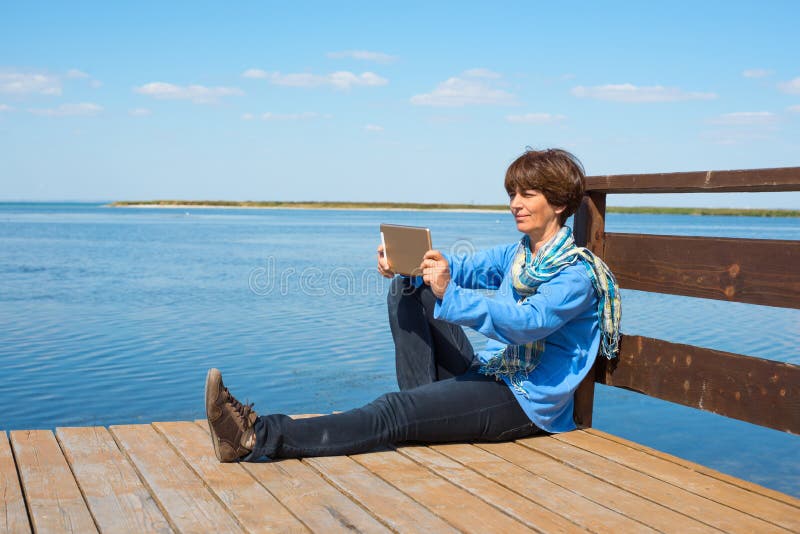 Happy woman is sitting on a wooden pier with tablet stock images Happy woman is sitting on a wooden pier with tablet stock images