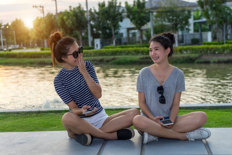 Happy women friends talking and laughing gossip holding mobile phone in a park outdoors with green background. .Two young Asian