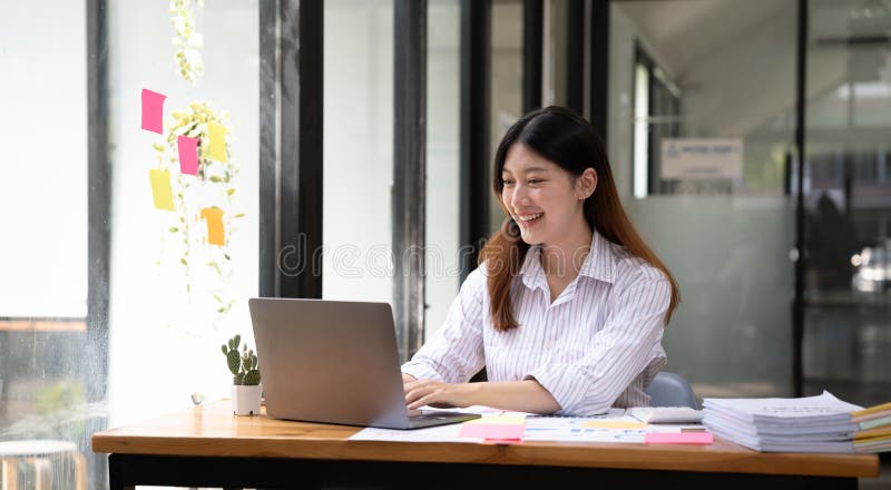 Happy young asian businesswoman sitting on her workplace in the office. Young woman working at laptop in the office.