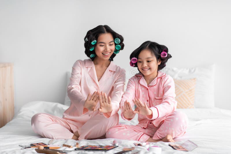 Happy young asian female and teenage girl with curlers looking at fresh nail polish sit on bed with cosmetics